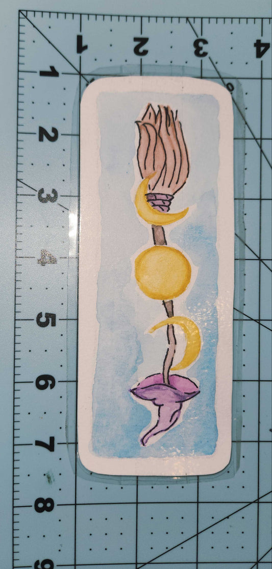 Card with a stylized illustration of a broom with yellow and purple elements on a blue background, placed on a cutting mat with measurement lines.