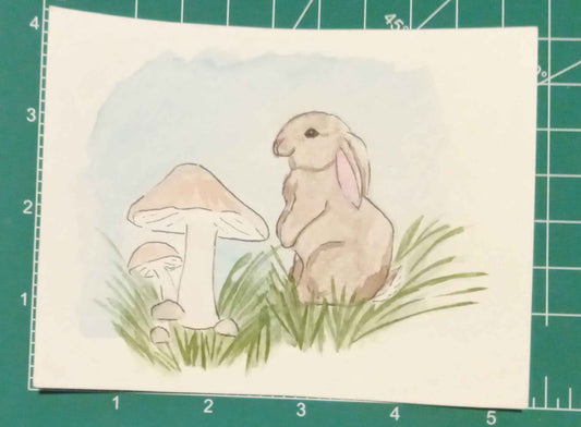 Card with a rabbit and mushroom illustration on a green cutting mat.