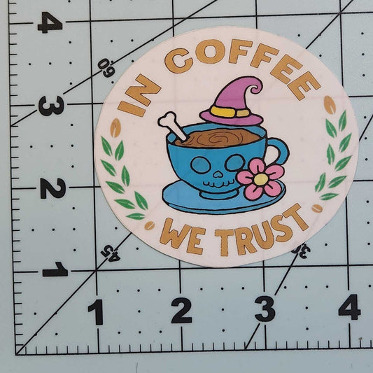 Circular sticker with cartoon character and text 'In Coffee We Trust' on a cutting mat with ruler.