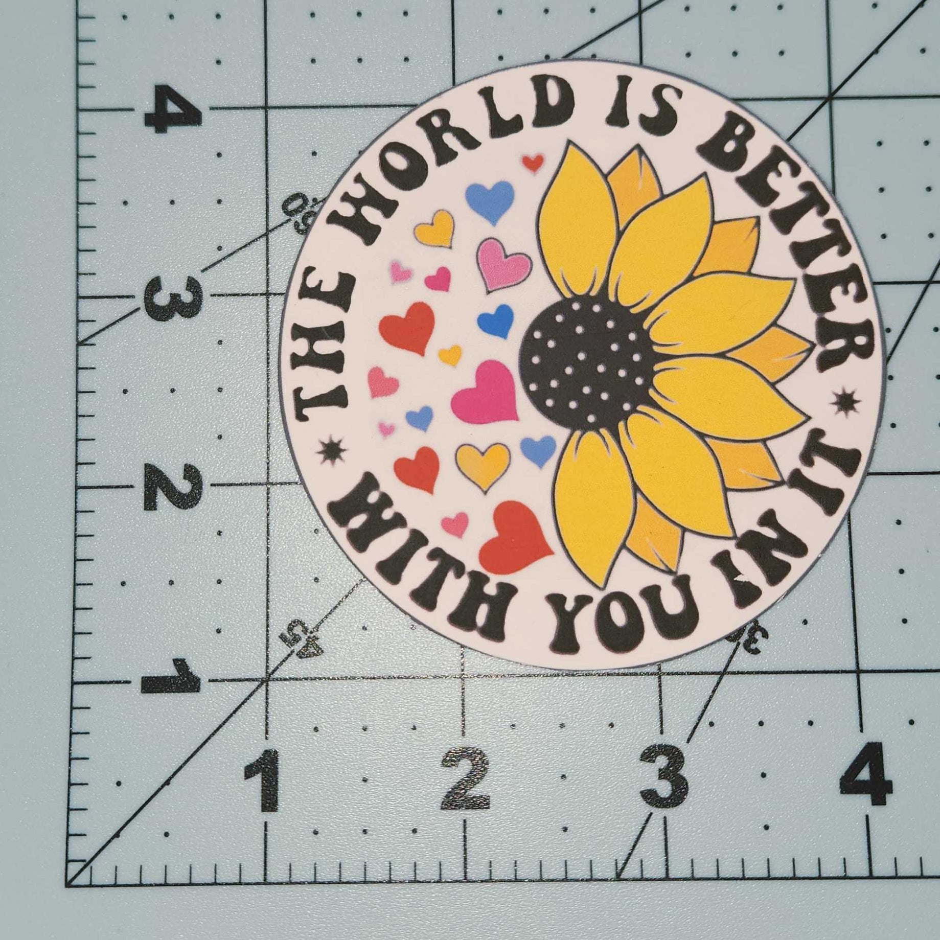 Sunflower sticker with text 'The world is better with you in it' on a cutting mat.