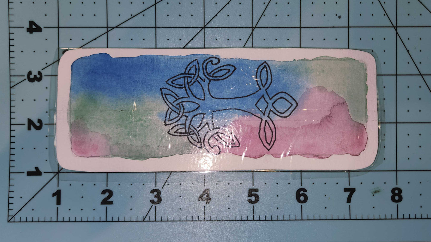 Decorative card with Celtic knot design on a watercolor background, placed on a cutting mat with measurements.