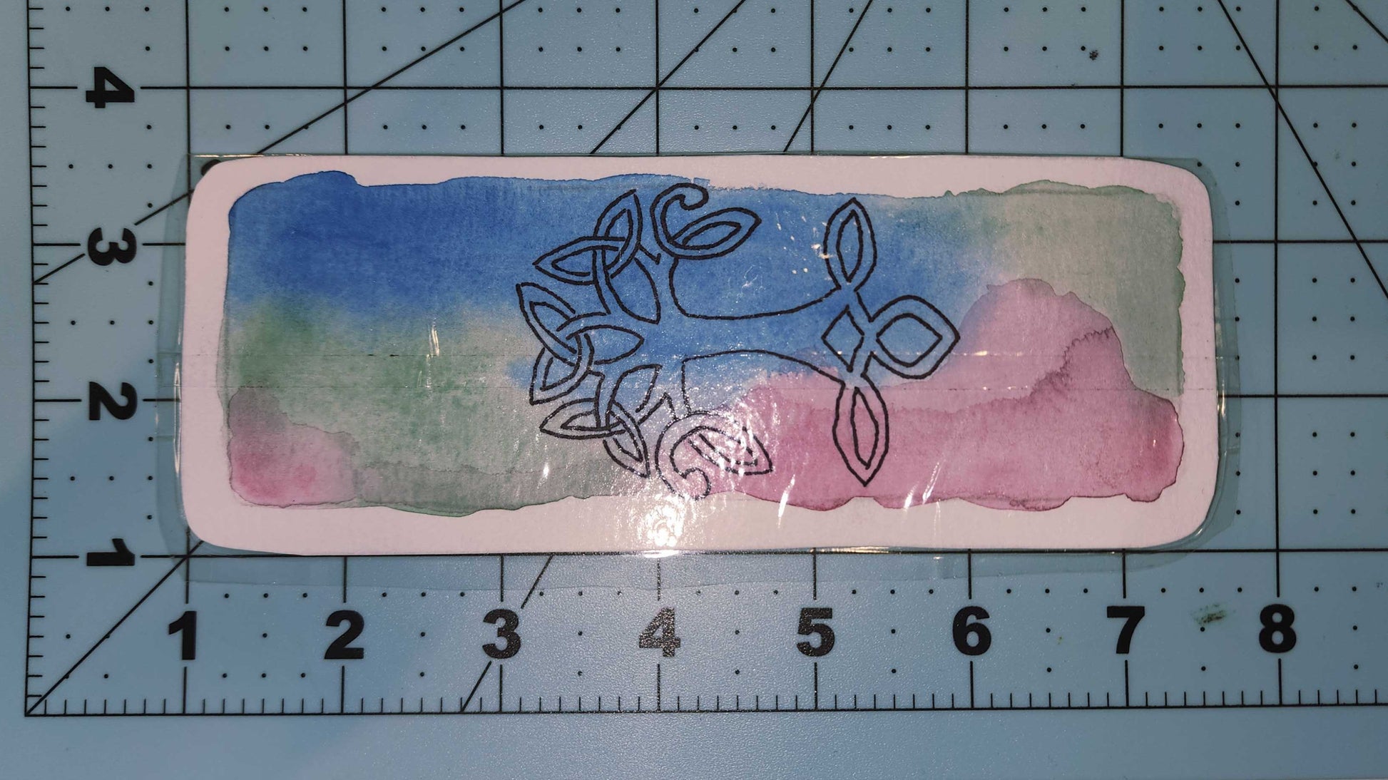 Decorative card with Celtic knot design on a watercolor background, placed on a cutting mat with measurements.