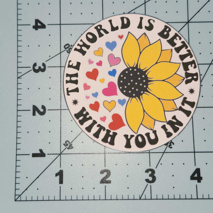 Sunflower sticker with text 'The world is better with you in it' on a cutting mat.